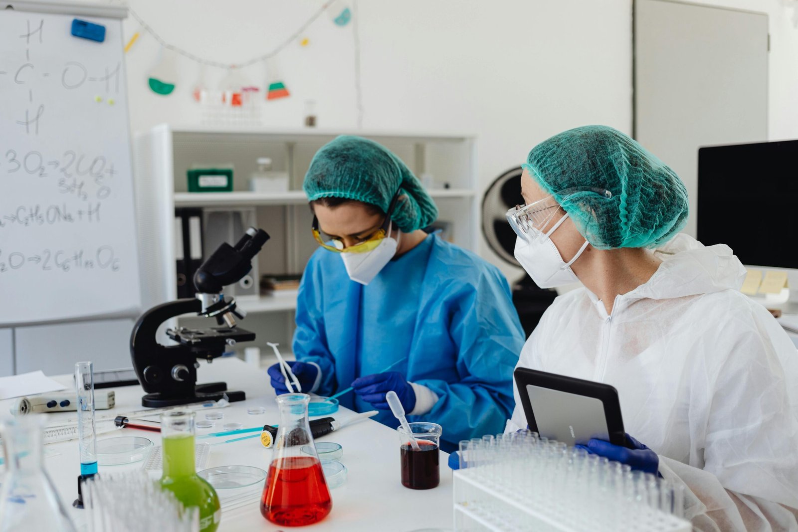 Two female scientists wearing protective gear conducting research in a lab with chemical samples and a microscope.